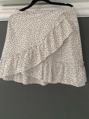 Vit prickig volangkjol från SHEIN - Superfin vit kortkjol från SHEIN med svarta prickar och volangdetaljer. Kjolen har omlottkänsla framtill och är perfekt för dig som gillar en söt och trendig look. Lätt och luftig, passar perfekt till varmare dagar.