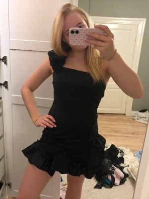 Svart oneshoulder volangklänning - Supersnygg svart kortklänning med volangdetaljer och oneshoulder-design. Klänningen har en tight passform 