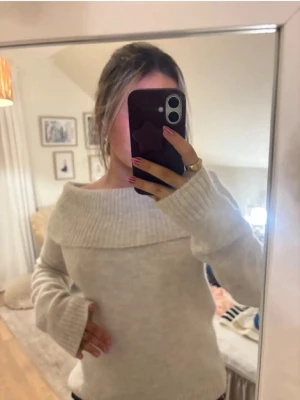 Offshoulder stickad tröja i beige - Supermysig stickad offshoulder-tröja i beige med bred ribbad kant upptill och långa ärmar. Perfekt för dig som vill ha en chill men ändå snygg look. Passar till jeans eller kjol och är lätt att styla med smycken.