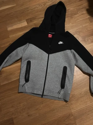 Grå svart Nike tech hoodie med dragkedja storlek S - Säljer en grå svårt Nike Tech Hoodie med dragkedja i storlek S, tröjan är nästan aldrig använd. Pris kan diskuteras