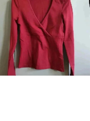 VinRöd omlottopp med v-ringning - Snygg röd omlottopp med djup v-ringning och långa ärmar. Toppen har en figurnära passform och är tillverkad i mjuk bomull som känns skön mot huden. Perfekt att styla med jeans eller kjol för en trendig look.