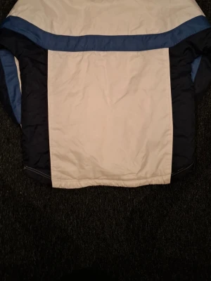 Retro pufferjacka från FILA i blått och vitt - Säljer en retro pufferjacka från FILA i vitt, mörkblått och blått. Jackan har klassiska färgblock, ribbade muddar och dragkedja med knappar. Materialet är nylon med polyesterfoder, perfekt för kalla dagar. Stor logga på bröstet och schysst vintagekänsla.