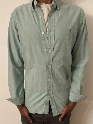 Grön skjorta från Polo Ralph Lauren - Klassisk grön skjorta från Polo Ralph Lauren med button-down krage och broderad logga på bröstet. Skjortan har långa ärmar och knappar framtill. Perfekt till jeans eller chinos för en clean look.