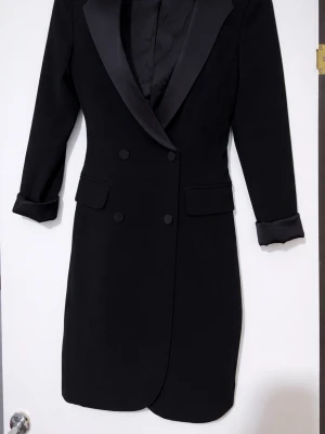 Svart kavaj med satindetaljer - Snygg kavaj med breda slag i satin och dubbelknäppning framtill. Kavajen har två fickor med lock. Perfekt för dig som vill ha en stilren och elegant look. (Mer som en S)