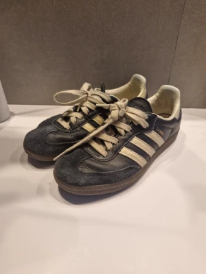 Adidas Samba sneakers mörkblå/vit - Klassiska Adidas Samba sneakers i svart skinn med vita detaljer och bruna gummisulor. Skorna har de ikoniska tre ränderna på sidan, snörning och låg profil. Perfekta för dig som gillar retrostil och streetwear. Det tillkommer också mörkblå snören som man kan byta som man ser på sista bilden. Om du har frågor är det bara att skriva och pruta