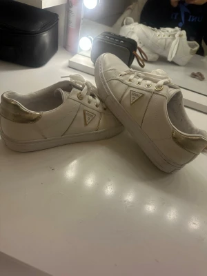 Vita och guldiga sneakers från Guess - Snygga vita sneakers från Guess med gulddetaljer och logga på sidan. Skorna har vita snören, guldfärgade öljetter och en metallic guldhäl. Tillverkade i syntetmaterial med platt sula och rund tå. Perfekta för dig som vill ha en clean men ändå lyxig look.
