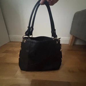 Svart handväska med gulddetaljer - Snygg svart handväska i skinnimitation med guldfärgade detaljer och kedjor. Väskan har dubbla handtag, dekorativ tofs och flera fack med dragkedja både inuti och utanpå. Perfekt storlek för allt du behöver ha med dig och riktigt trendig look.