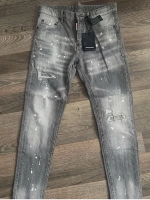 Grå  jeans från Dsquared2 - Säljer ett par grå jeans från Dsquared2 med coolt blekt tvättad look och vita färgstänk. Jeansen har slitna detaljer vid knäna och klassisk femficksdesign. Materialet är jeans med stretch för skön passform. Perfekta för dig som vill sticka ut lite extra.