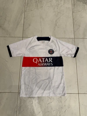 PSG matchtröja  - Snygg PSG matchtröja i vitt med marinblå och röda detaljer samt klubbmärke på bröstet. Tröjan har korta ärmar, rund hals och är tillverkad i lätt polyester. Har endast testat på. 