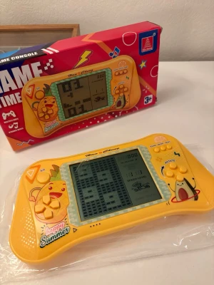 Han Cheng Handheld Game Console - Färgglad retro LCD-spelkonsol från Han Cheng med flera klassiska spel som Tetris. Stor skärm, enkla kontroller och rolig design med fruktmotiv. Passar barn från 3 år. Konsolen är i mycket fint skick och levereras med originalkartong.