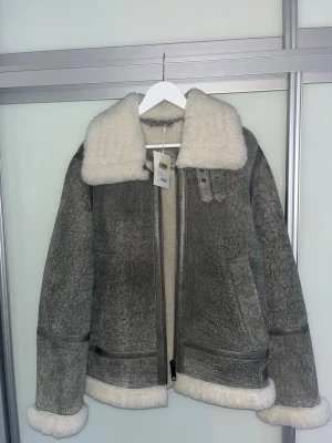 Grå shearling från GANT, storlek M - GANT shearlingjacka i äkta lammläder Snygg och tidlös shearlingjacka från GANT, Utsida i mjukt läder/mocka och insida i varm, naturlig ull – perfekt för kalla dagar. Klassisk modell med kraftig dragkedja, bred shearlingkrage och justerbara spännen i midjan. Äkta shearling Väldigt varm och bekväm Färg: grå / desert brown Storlek: M Skick: mycket fint / nyskick (med tag kvar) Nypris: 20 0000 En exklusiv jacka av hög kvalitet som håller i många år. Passar både vardag och mer uppklädd