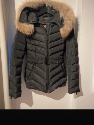 Svart pufferjacka Hollies med päls - Säljer en svart pufferjacka från Hollies med avtagbar huva och fluffig beige päls. Jackan har bälte i midjan, dragkedja framtill och två fickor med dragkedja. Perfekt för kalla dagar och riktigt snygg siluett.