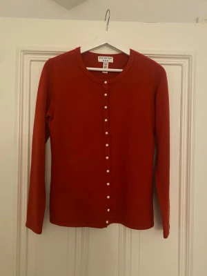 Röd kofta med pärlknappar Covington - Snygg röd kofta från Covington i storlek S. Koftan har rund halsringning och pärlknappar längs hela framsidan. Tillverkad i mjukt akrylmaterial och har långa ärmar. Perfekt för att addera lite färg till din outfit!