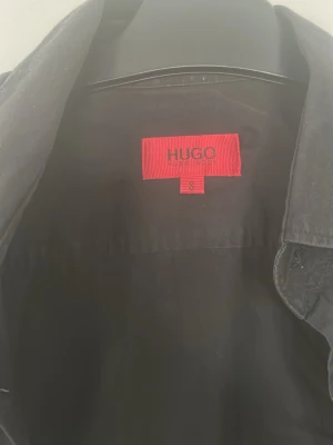 Svart skjorta från HUGO - Klassisk svart skjorta från HUGO med knappar framtill och lång ärm. Skjortan har en stilren design och mjukt material som känns skönt mot huden. Perfekt för dig som gillar minimalistisk och clean stil.