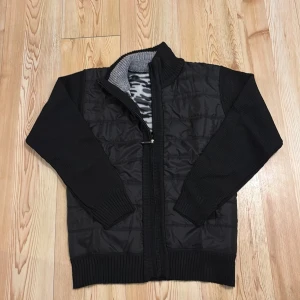 Svart quiltad jacka med dragkedja - Säljer en svart quiltad jacka med dragkedja och ribbade muddar. Insidan har ett mönstrat foder i grått och svart. Perfekt för kyliga dagar!