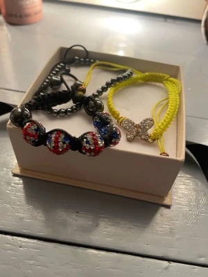 Armband - Säljer tre snygga armband – ett svart med glittrande pärlor i rött, blått och silver, och ett gult med en gnistrande fjäril i mitten, och ett silver med silver pärlor. Alla har justerbara band i flätad stil och passar perfekt för att ge outfiten lite extra bling. Kontakta om du är intresserad av ett armband istället för alla🤍