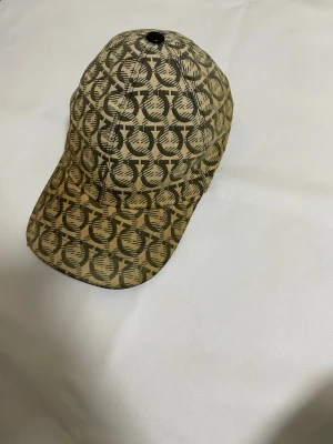 Beige mönstrad keps från Ferragamo - Säljer en beige keps från Salvatore Ferragamo med ett unikt, grafiskt mönster i mörkbrunt. Kepsen har böjd skärm och justerbar rem bak i svart. Tillverkad i Italien och har en lyxig känsla med snygg design som sticker ut.