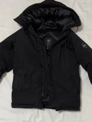 Svart dunjacka från Canada Goose - Svart dunjacka från Canada Goose med huva och quiltad insida. Jackan har dragkedja och knappar framtill, två sidofickor och en patch med logga på ärmen. Yttermaterialet är polyester och bomull, fodret är nylon och fyllningen består av vitt dun och fjäder.