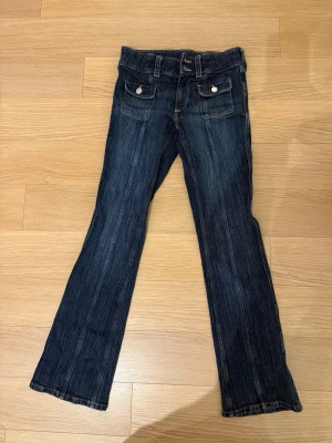 Bootcut jeans med låg midja från H&M  - Mörkblå bootcut jeans från H&M med låg midja och snygga fickor med knappar framtill. Jeansen har ett coolt randigt mönster i denim och klassiska bälteshällor. Perfekt för dig som gillar en avslappnad och trendig stil.