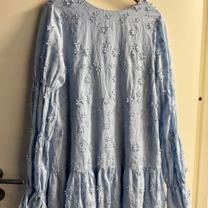 Nakd klänning - Supersöt ljusblå klänning med broderade blommor över tyget. Klänningen har långärmade volangärmar och en djup rygg med knytband. Den är kort i modellen och har en flowy, avslappnad passform. Perfekt för dig som gillar romantiska detaljer och mjuka färger. Storlek 36🩵