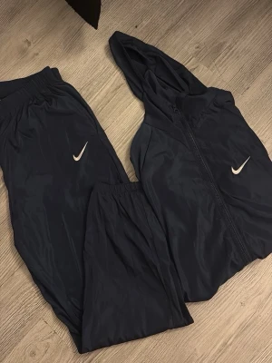 Svart Nike träningsset med hoodie - Snyggt svart träningsset från Nike med matchande hoodie och byxor. Båda delarna har den klassiska vita Nike-loggan. Setet är gjort i lätt syntetmaterial som är perfekt för träning och sport. Hoodien har dragkedja och byxorna har resår i midjan.
