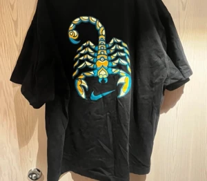 Svart Nike SB t-shirt med skorpiontryck - Svart t-shirt från Nike SB med ett färgstarkt skorpiontryck i blått och gult på ryggen samt liten Nike SB-logga i blått på bröstet. Klassisk rund hals och korta ärmar. Materialet är mjuk bomull som känns skönt mot huden.