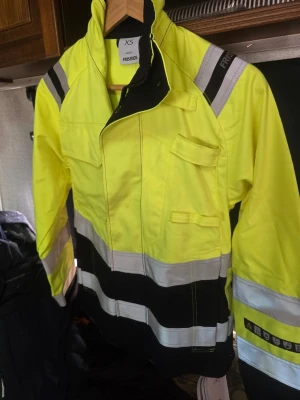 Gul vindjacka från Fristads XS - Säljer en gul vindjacka från Fristads i storlek XS. Jackan har svarta och vita reflexdetaljer, flera fickor och dragkedja framtill. Perfekt för dig som vill synas och ha en praktisk jacka med cool design och funktionella detaljer.
