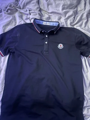 Svart Moncler pikétröja  - Säljer en svart pikétröja från Moncler i storlek M. Tröjan har klassisk krage med röd och vit randig detalj, knäppning med tre knappar och Moncler-logga broderad på bröstet. Materialet är mjuk bomull och modellen har korta ärmar med matchande rand på ärmslut. Liten i storleken passar xs/s