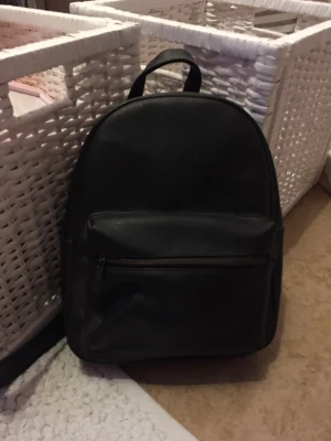 Svart mini ryggsäck i skinnimitation - En stilren svart mini ryggsäck med enkel design och praktisk ytterficka med dragkedja. Väskan har ett stort huvudfack och är tillverkad i skinnimitation. Perfekt för dig som vill ha en clean look och plats för allt du behöver under dagen. 