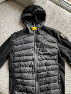 Svart pufferjacka Parajumpers S - Svart pufferjacka från Parajumpers med huva och gul detalj vid halsen. Jackan har quiltad framsida, dragkedja hela vägen och en bröstficka med dragkedja. Insidan har flera fickor och orange detaljer. Perfekt för kyliga dagar.