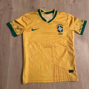Brasilien fotbollströja - Säljer en officiell Brasilien fotbollströja från Nike i gul med gröna detaljer på krage och ärmslut. Tröjan har tryckt Brasilien-emblem och Nike-logga framtill samt ett subtilt motiv av Kristusstatyn både fram och bak. Perfekt för dig som älskar fotboll och Brasilien.