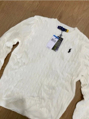 Vit stickad tröja från Polo Ralph Lauren - Snygg vit stickad tröja från Polo Ralph Lauren med klassisk kabelstickning och broderad logga på bröstet. Rund hals och långa ärmar, perfekt för att hålla sig varm och ändå se fräsch ut. Passar dig som gillar stilrena och tidlösa plagg.