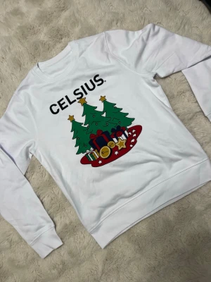 Vit jultröja från Celsius med tryck. Kolla beskrivning! - Säljer en vit sweatshirt från Celsius med stort juligt tryck framtill. Motivet har tre gröna julgranar med stjärnor, presenter, polkagrisar och Celsius-detaljer. Tröjan har rund hals och långa ärmar, perfekt för dig som vill sticka ut under julen. Har storlek S och XL skriv till mig vilken storlek du vill ha! Skickas samma dag!