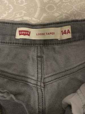 Grå Levi's Loose Taper jeans 14A - Säljer ett par grå jeans från Levi's i modellen Loose Taper. De har en avslappnad passform med avsmalnande ben och klassiska fem fickor. Jeansen är tillverkade i slitstarkt denim och har tydliga sömmar för en cool look.