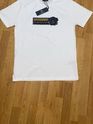 Helt ny! T-shirt - Snygg vit t-shirt i 100% bomull. Framsidan har en broderad logga i svart, gult och grått med Medusa-detalj. Klassisk passform och korta ärmar, perfekt till jeans eller shorts. Tillverkad i Italien.