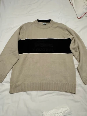 Beige stickad tröja från Lacoste - Säljer en beige stickad tröja från Lacoste med svart bred rand över bröstet och logotyp i mitten. Tröjan har rund hals och ribbade muddar vid ärmslut och nederkant. Perfekt för dig som gillar klassisk stil med en modern touch.