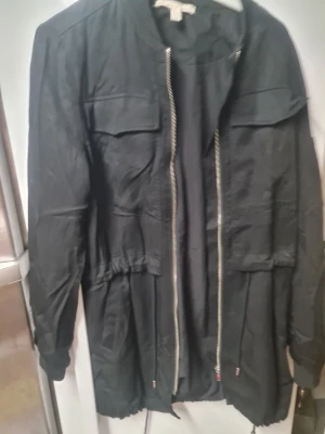 Svart parkajacka från trf_outerwear - Svart parkajacka med dragkedja framtill, stora fickor och snörning i midjan och nederkant. Jackan har lång ärm med extra ficka och dragkedja på armen. Materialet är troligtvis polyester och jackan har en loose passform. Perfekt för dig som gillar streetstyle.