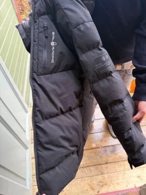 Svart pufferjacka från Sail Racing - Säljer en svart pufferjacka från Sail Racing med Sail Antarctica-märke på ärmen och dragkedja framtill. Jackan har en hög krage, flera fickor med dragkedja och är perfekt för kalla dagar. Snygg och stilren design som håller värmen.