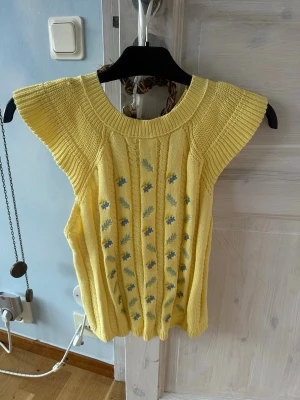 Gul stickad ärmlös tröja med blommor - Supersöt ljusgul stickad tröja med bred ribbad krage och ärmlös design. Finns någon liten söm som gått upp, se axeln. Framsidan har broderade små blommor i blått, lila och gult samt gröna blad. Tröjan har ett mjukt, kabelstickat mönster och känns perfekt för vår och sommar. Passar dig som gillar vintagevibbar och färg!