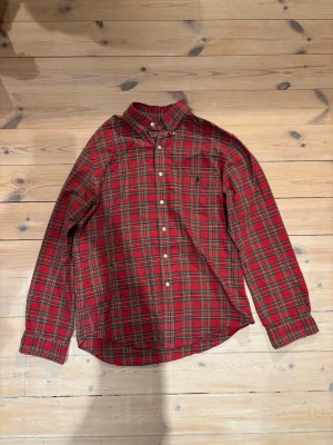 Rutig röd skjorta från Ralph Lauren - Säljer en slim fit skjorta från Ralph Lauren i storlek L. Skjortan är röd med klassiskt rutigt mönster i grönt, blått och gult. Den har långa ärmar, knappar framtill och krage. Materialet är mjuk bomull, perfekt för dig som gillar preppy stil.