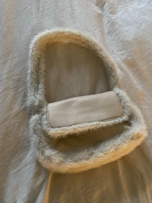 Beige axelväska med fuskpäls - Säljer en beige axelväska med mjuka fuskpälsdetaljer runt kanterna och på axelremmen. Väskan har en enkel, rektangulär form och är tillverkad i syntetmaterial med pälsimitation. Perfekt för dig som gillar unika accessoarer med cozy vibe.