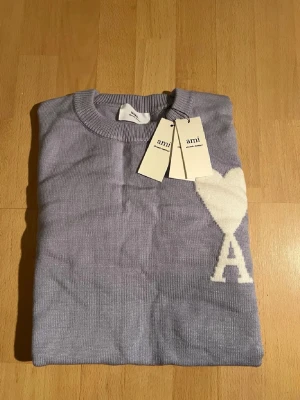 Ljuslila stickad tröja från Ami - Snygg ljuslila stickad tröja från Ami med ett stort vitt hjärta och bokstaven A på bröstet. Rund halsringning och ribbade muddar. Perfekt för dig som vill ha en trendig och stilren look med en unik detalj.
