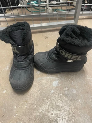Svarta vinterboots från Sorel - Svarta boots från Sorel med fluffigt foder och bred rem med logga över foten. Skorna har rund tå, platt sula och är tillverkade i syntetmaterial med extra vaddering för värme. Perfekta för kalla dagar och snö, med robust design och hög komfort.
