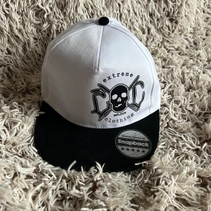 Vit och svart snapback från Extreme Clothing - Säljer en snapback-keps från Extreme Clothing i vit och svart med döskalletryck framtill. Kepsen har justerbar bakdel och klassisk platt skärm. Materialet är bomull och polyester, perfekt för att ge din outfit en edgy vibe.