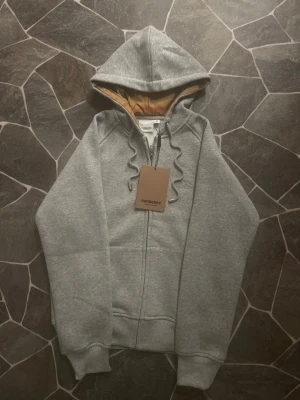 Grå hoodie från Burberry med dragkedja - Snygg grå hoodie från Burberry med klassisk logga broderad på bröstet och ikoniskt rutigt foder i huvan. Tröjan har snörning vid huvan, ribbade muddar och en rejäl dragkedja framtill. Perfekt för dig som gillar stilrena och lyxiga streetwear-plagg. Plagget sitter som S - M.