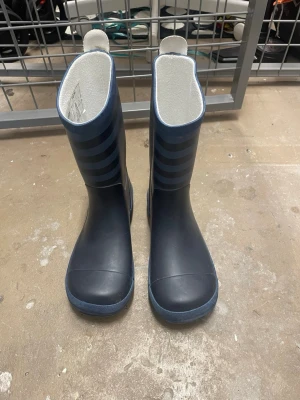 Mörkblå randiga stövlar - Säljer ett par mörkblå stövlar med snygga horisontella ränder i en lite ljusare blå ton. Insidan är vit och mjuk, och stövlarna har rund tå och platt sula. Perfekta för regniga dagar och gjorda i slitstark syntet. Enkla att dra på med praktisk flik upptill.