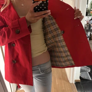 Röd trenchcoat med bälte - Nypris: Ungefär 2 500 - 3 500 kr.                     Snygg röd trenchcoat med klassisk knäppning och bälte i midjan. Jackan har stora knappar, fickor på sidorna och rutigt foder på insidan. Perfekt för dig som vill sticka ut med en färgstark look. Passar till både jeans och kjol. Lite nopprig på insidan men ananrs som ny.          