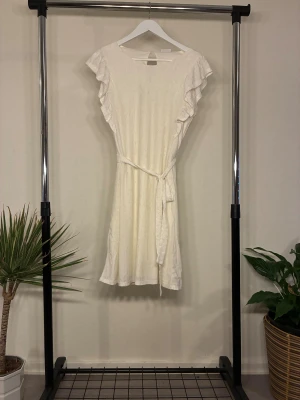 Vit kortklänning med volang från Vila - Supersöt vit kortklänning från Vila med volangdetaljer på ärmarna och knytband i midjan. Klänningen har ett diskret hålmönster och är tillverkad i ett mjukt material som känns luftigt och bekvämt.  Följer kroppens former och passar även M för mer slimmad look. Strolek L helt oanvänd. 