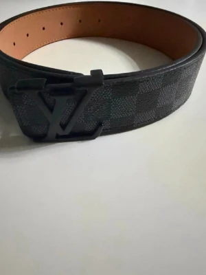 L&V bälte - Snyggt bälte med klassiskt rutigt mönster i svart och grått. Bältet har en stor logga i svart metall som spänne och insidan är i ljusbrunt skinn. Perfekt för att lyfta din outfit med en lyxig touch.