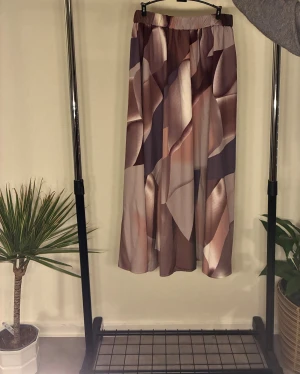 Mönstrad långkjol i satinlook - En flowy långkjol med abstrakt mönster i beige, brun och lila toner. Kjolen har ett glansigt material som ger en lyxig känsla. Storlek M, men passar även S då det beror på hur ni gillar er passform. 